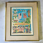 A framed folk art print depicting a street scene with a trolley, flags, and figures, mounted in a gold frame with a beige textured mat on a light marble surface.