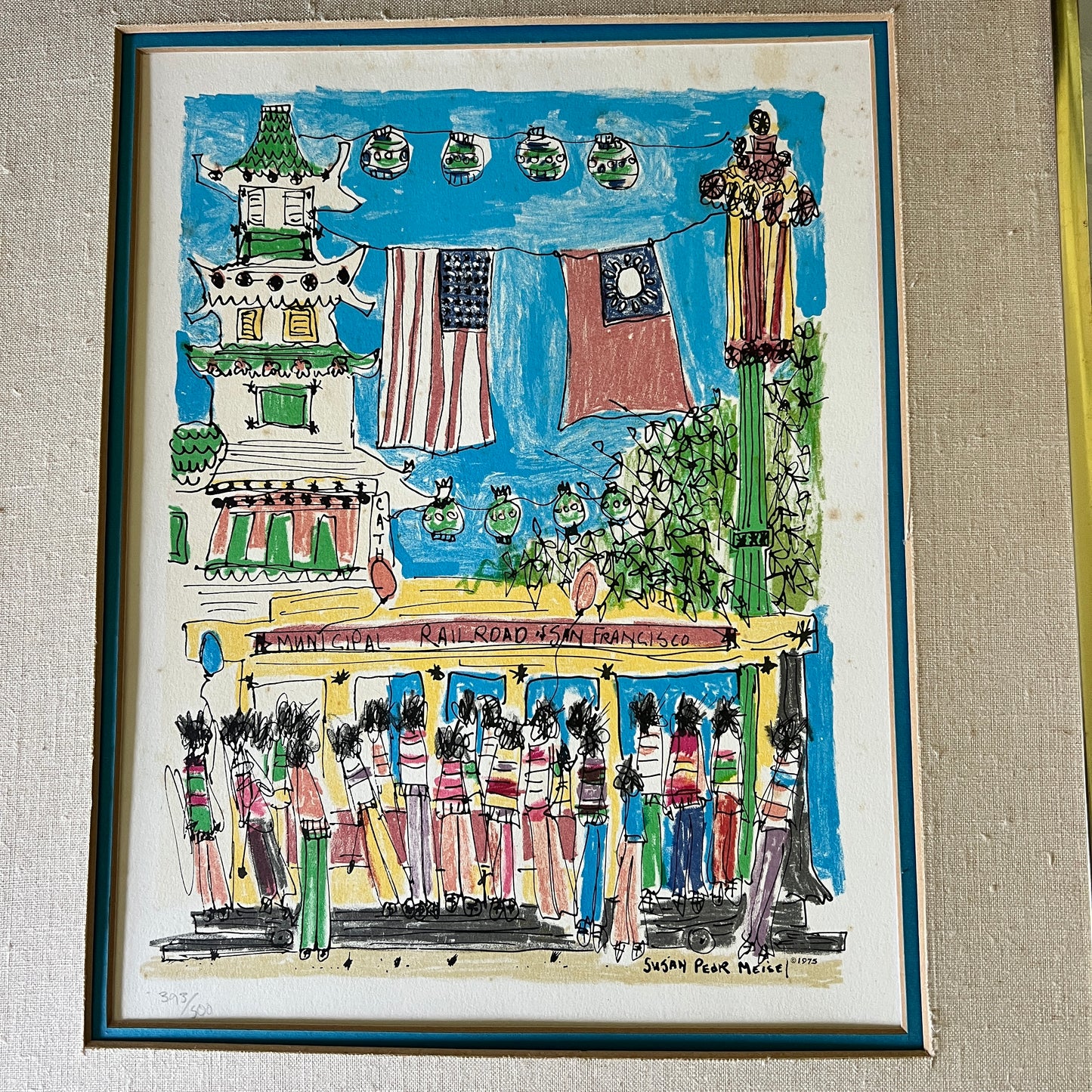 A framed folk art print depicting a street scene in San Francisco with a pagoda, hanging flags, a trolley, and figures walking along the street, mounted on a textured beige wall.