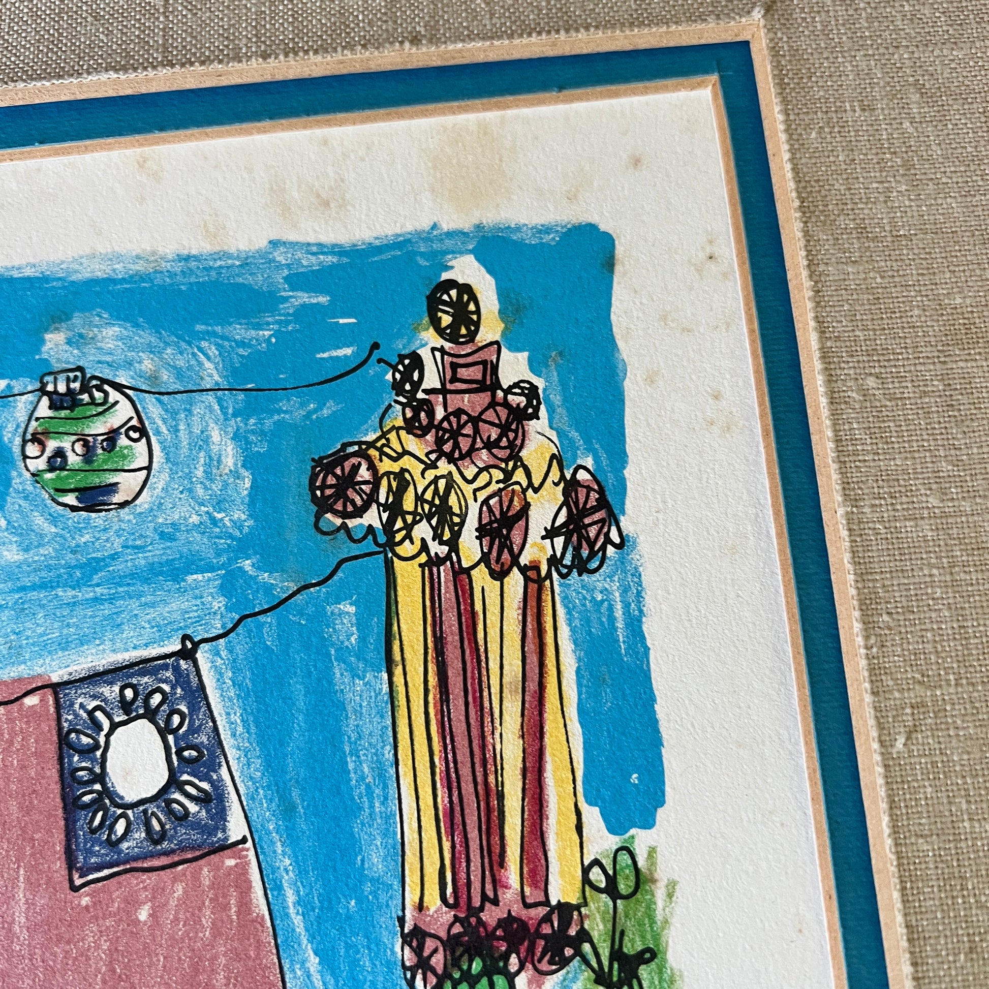 A close-up of a framed artwork on a textured beige surface. The artwork shows a childlike drawing of a tall figure in a yellow and red striped dress holding flowers, with a blue sky background, a hanging ornament, and a small blue-and-white flag.