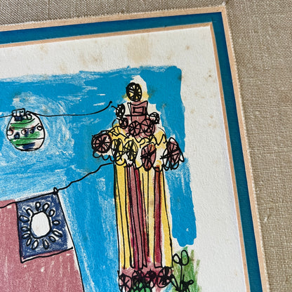 A close-up of a framed artwork on a textured beige surface. The artwork shows a childlike drawing of a tall figure in a yellow and red striped dress holding flowers, with a blue sky background, a hanging ornament, and a small blue-and-white flag.