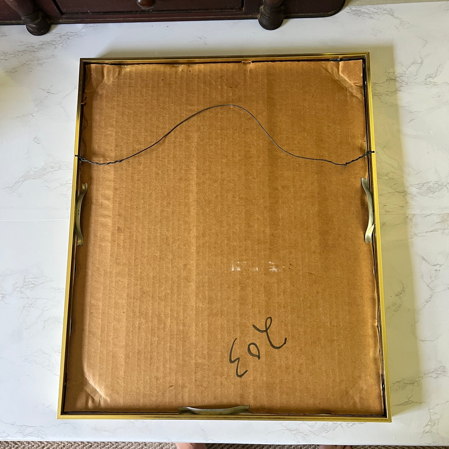 The back of a rectangular gold-framed picture frame, showing a brown cardboard backing with a hanging wire and handwritten numbers, placed on a white marble surface.