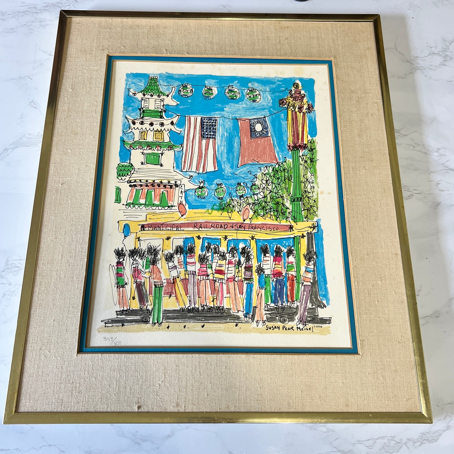 A framed art print depicting a colorful street scene with people, flags, and a building, mounted in a textured beige mat and a gold-toned frame on a light marble surface.