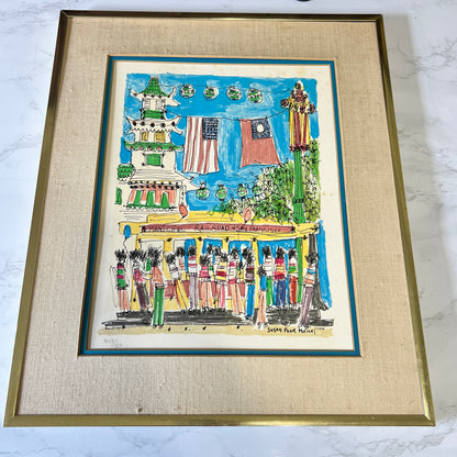 A framed art print depicting a colorful street scene with people, flags, and a building, mounted in a textured beige mat and a gold-toned frame on a light marble surface.