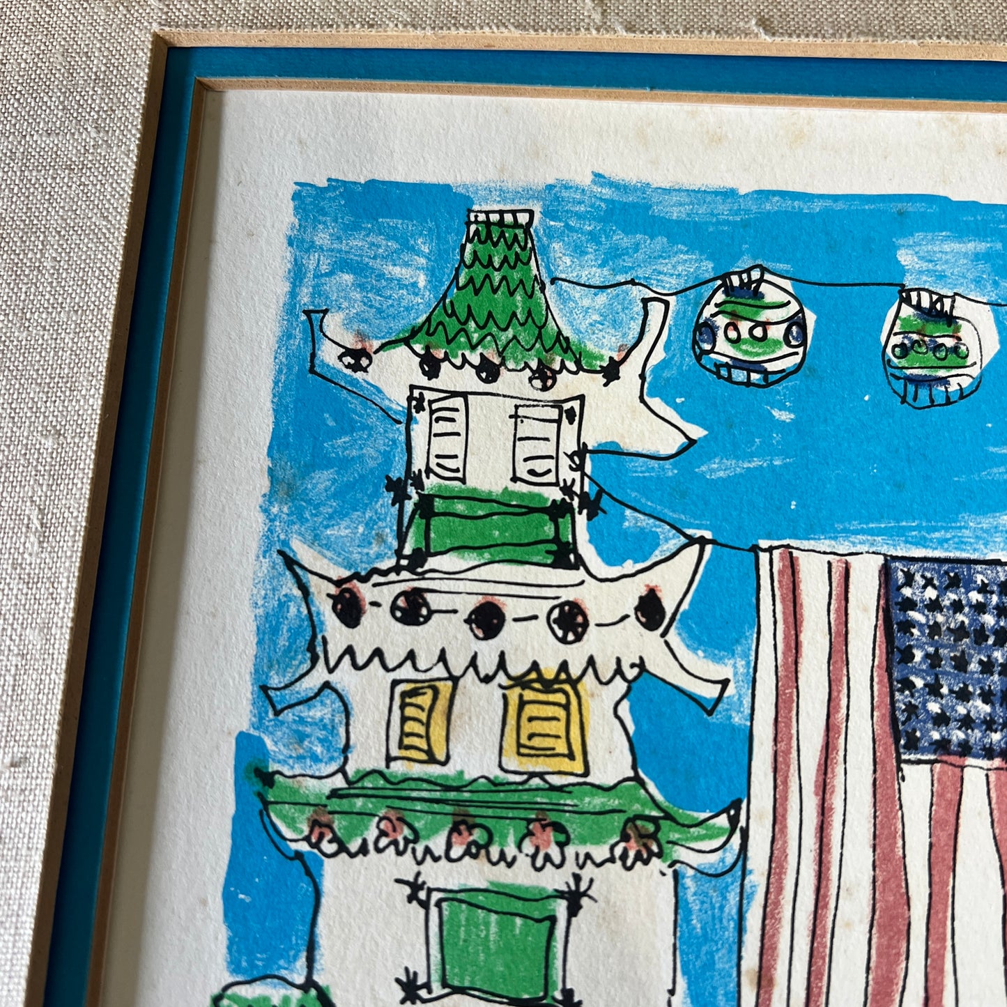 A framed hand-drawn children's art print featuring a green and white pagoda, a blue sky, and a partially visible American flag, shown in a close-up angled view.