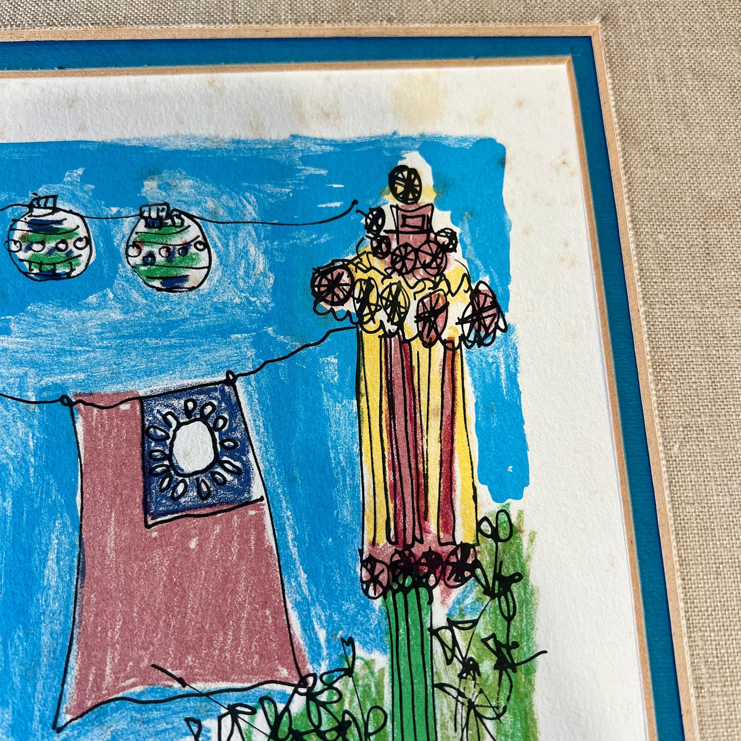A framed children's colored pencil drawing on textured paper, featuring a yellow striped Figure, a red and blue flag, and green Christmas ornaments against a blue sky, set in a blue and beige frame.