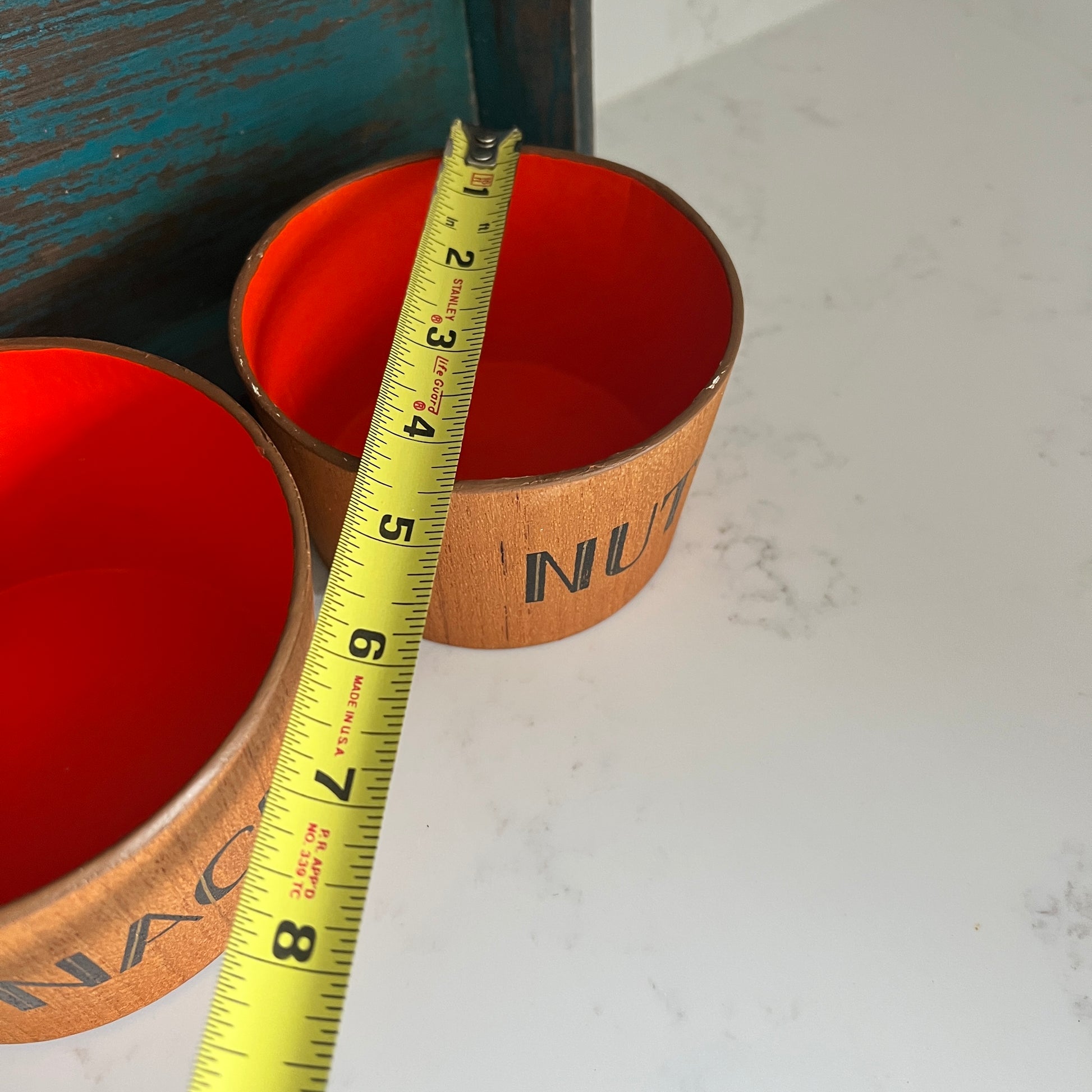 Two orange containers with wooden lids on a white surface, measured by a yellow tape measure.