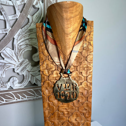 A carved wooden display bust wearing a patterned silk scarf and a bronze-toned medallion engraved with 'VPR 1870', set against a light gray wall with an ornate white carved panel in the background.