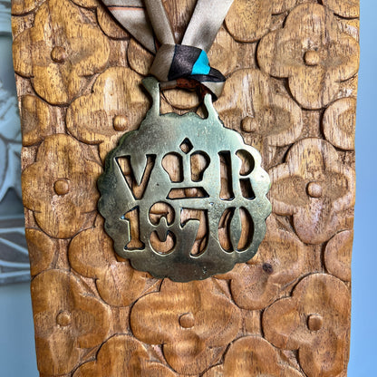 A close-up of a round metal pendant hanging from a ribbon, mounted on a carved wooden plaque with floral patterns. The pendant is aged bronze-toned with cutout lettering reading 'VRI 1870' and a crown motif.