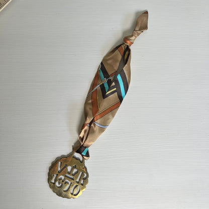 A vintage silk necklace with a gold-toned metal pendant medallion engraved with 'VIR 1350' and a crown, laid flat on a light gray textured surface.