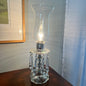 Clear glass hurricane lamp on a wooden surface with a light on inside, against a neutral wall.