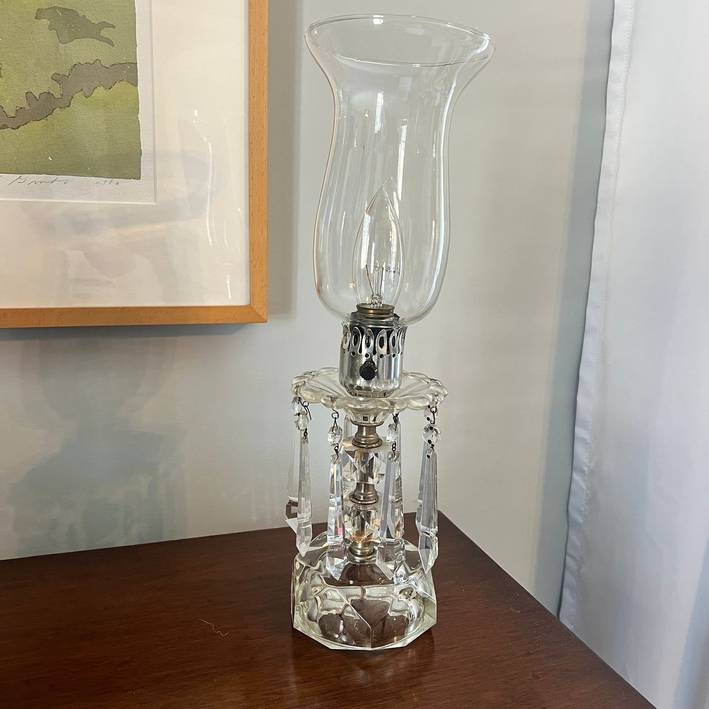 Clear glass hurricane lamp on a wooden surface with a framed picture in the background.