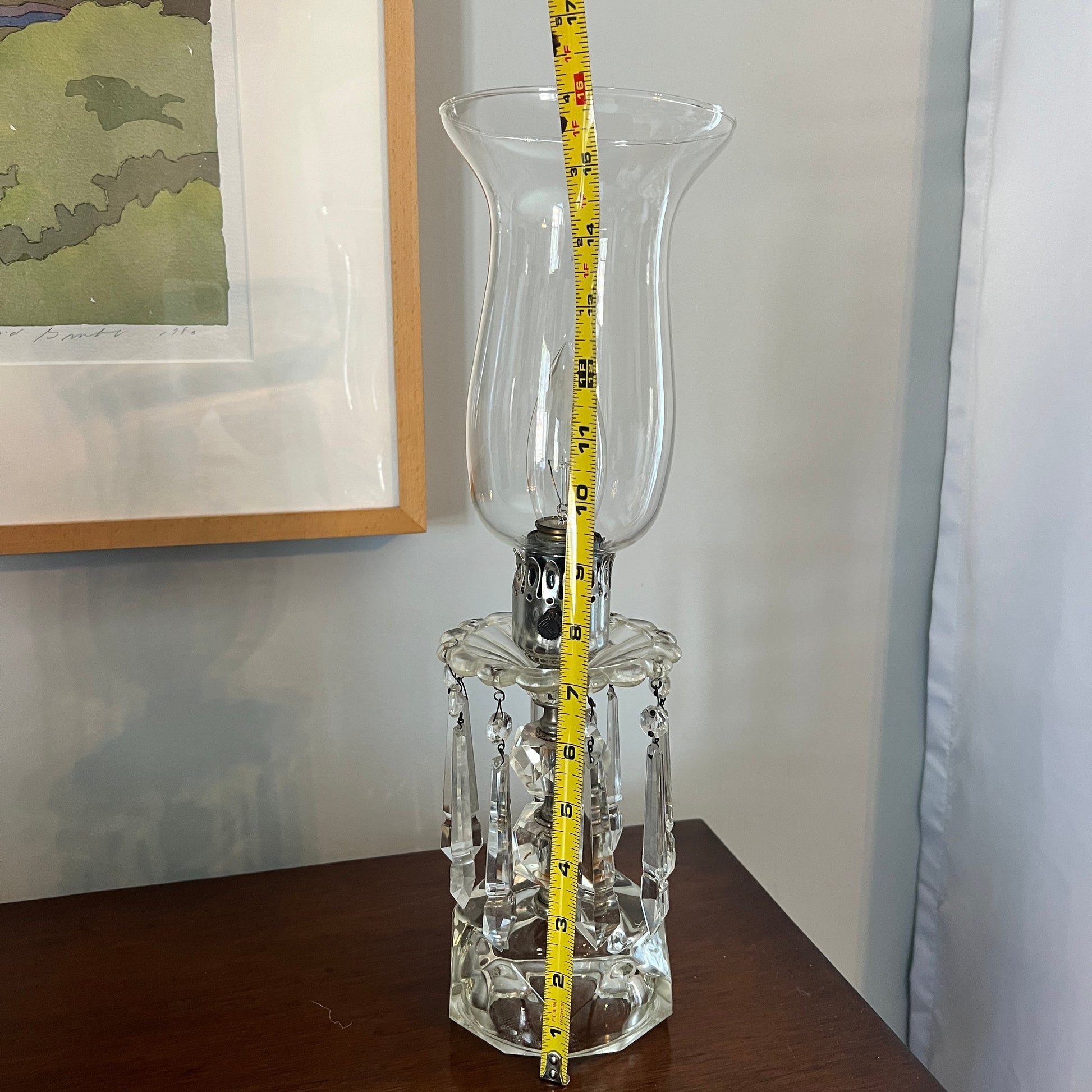 Clear glass hurricane lamp with a measuring tape for scale on a wooden surface.