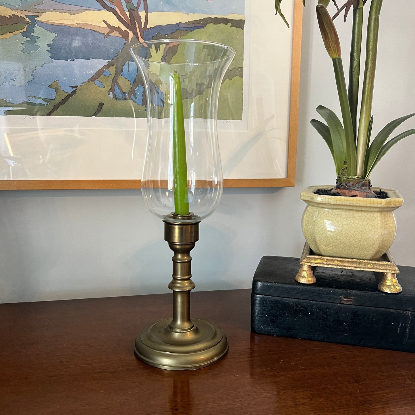 Decorative candle holder with a green candle on a wooden surface, next to a potted plant and framed artwork.