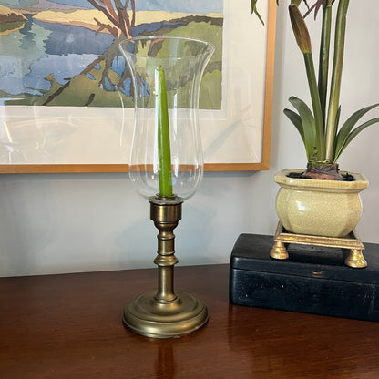 Decorative candle holder with a green candle on a wooden surface, next to a potted plant and framed artwork.