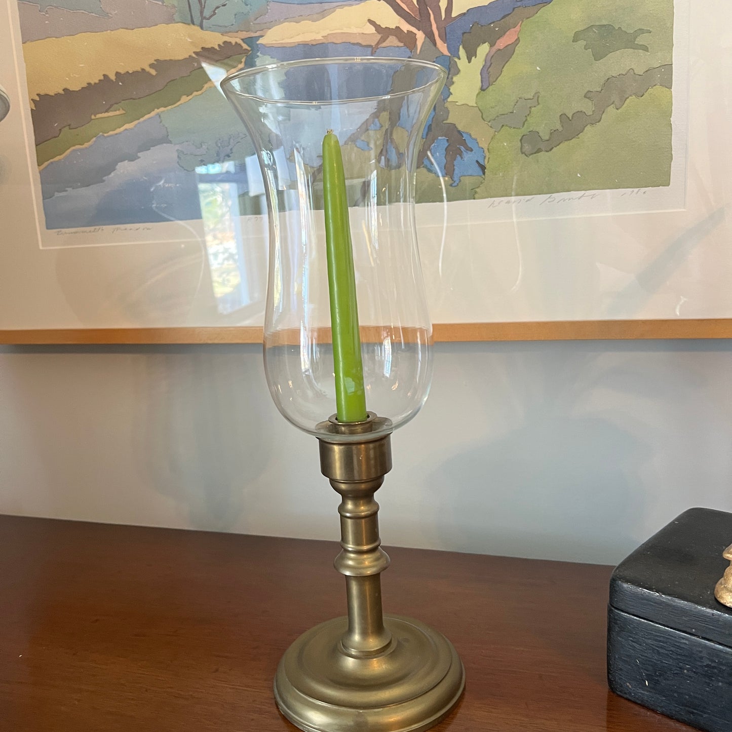 Brass candle holder with a green candle on a wooden surface, with a painting in the background.