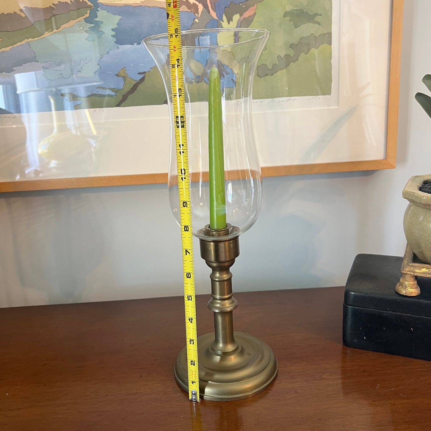 Brass candlestick holder with a green candle and a tape measure for scale, on a wooden surface with a painting in the background.