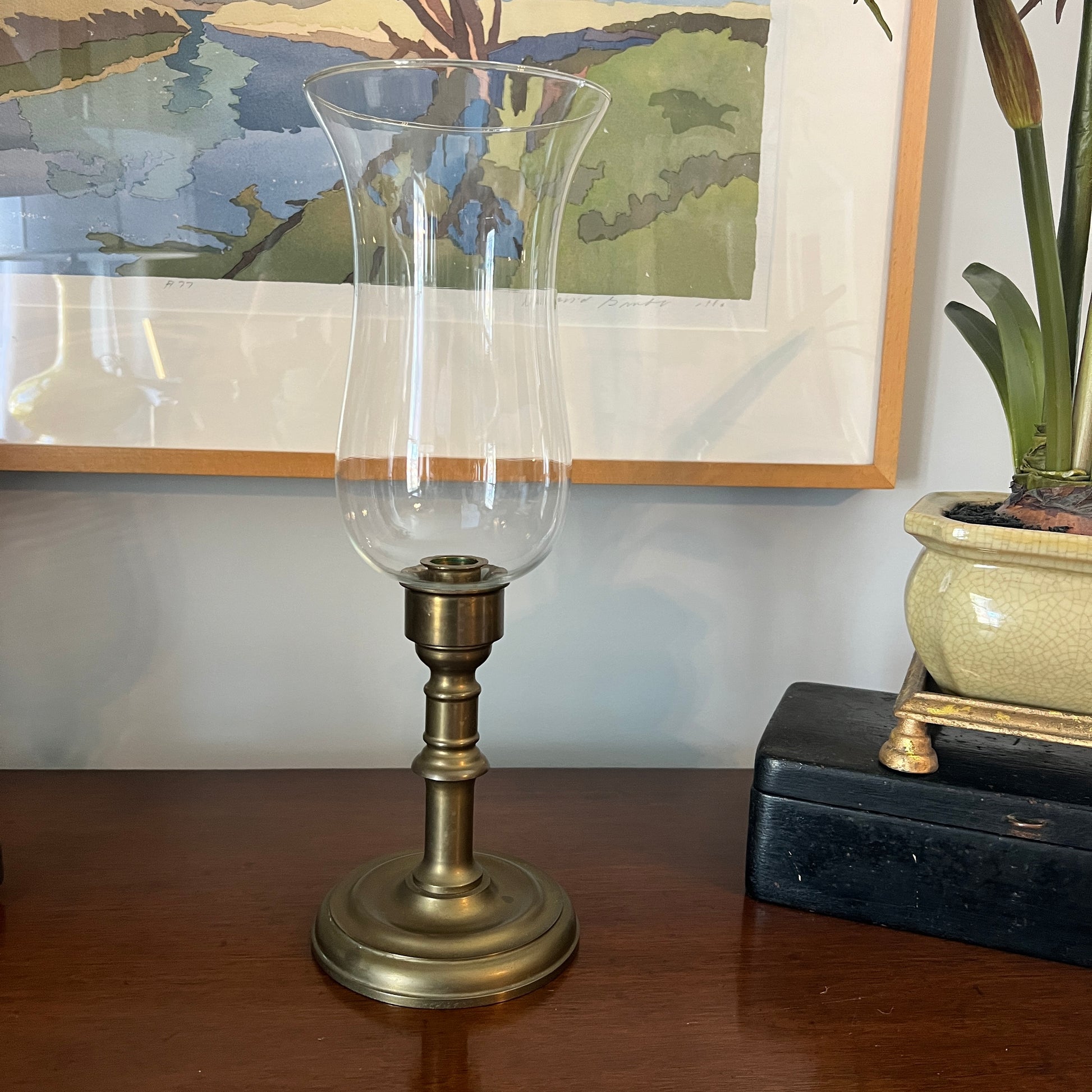 Decorative candlestick with clear glass shade and brass base on a wooden surface, with a painting and plant in the background.