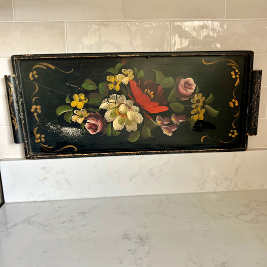 A rectangular black decorative tray with hand-painted floral motifs, including yellow flowers, white blossoms, pink roses, and a red flower, displayed on a white marble countertop against a tiled backsplash.