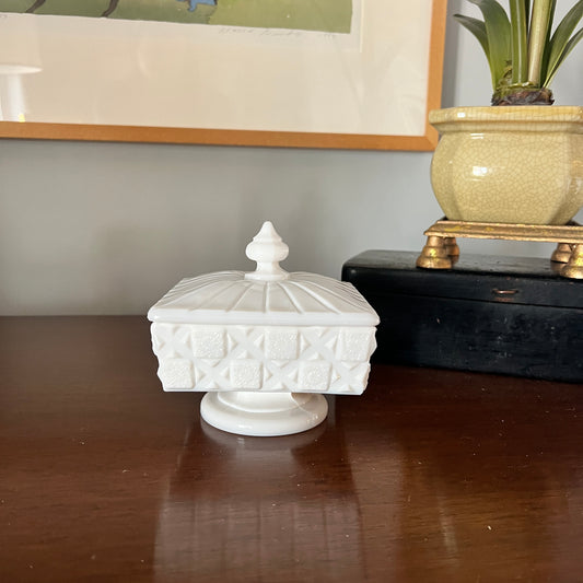 White textured lidded container on a wooden surface with a plant and framed picture in the background.