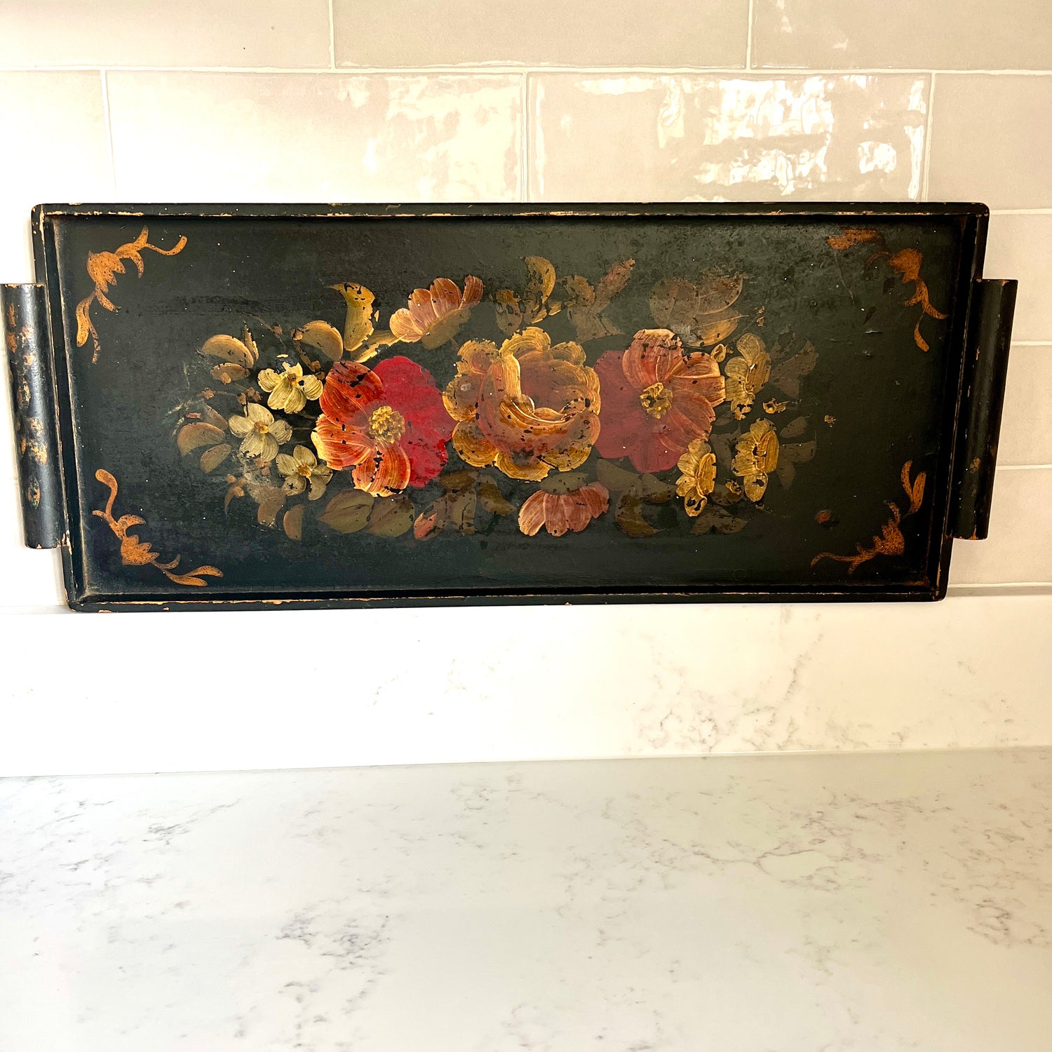 A rectangular black serving tray with a hand-painted floral design featuring red, orange, and yellow flowers, displayed on a white marble countertop against a white tiled wall.