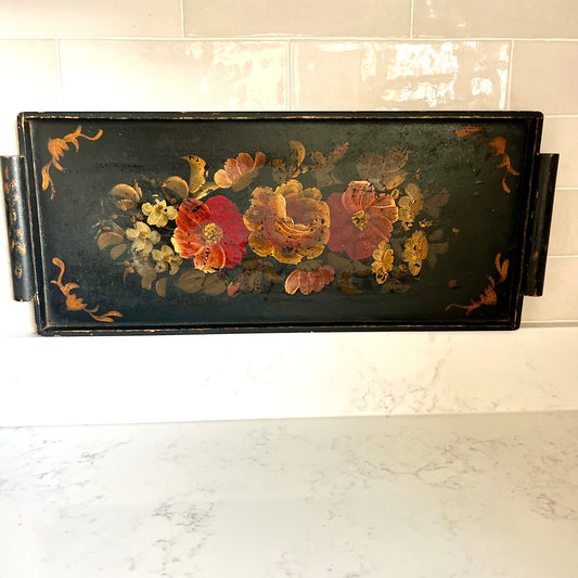 A rectangular black serving tray with a hand-painted floral design featuring red, orange, and yellow flowers, displayed on a white marble countertop against a white tiled wall.