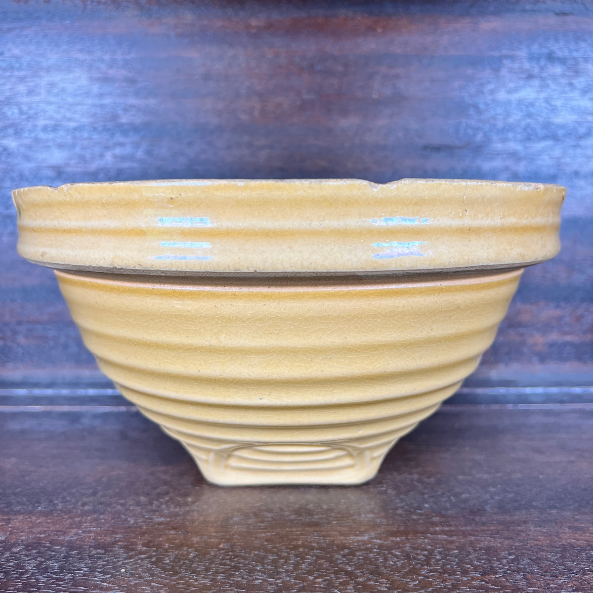 A handmade ceramic pottery with a warm yellow-beige glaze and horizontal ridges, set against a dark wooden background.