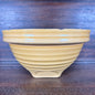 A handmade ceramic pottery with a warm yellow-beige glaze and horizontal ridges, set against a dark wooden background.