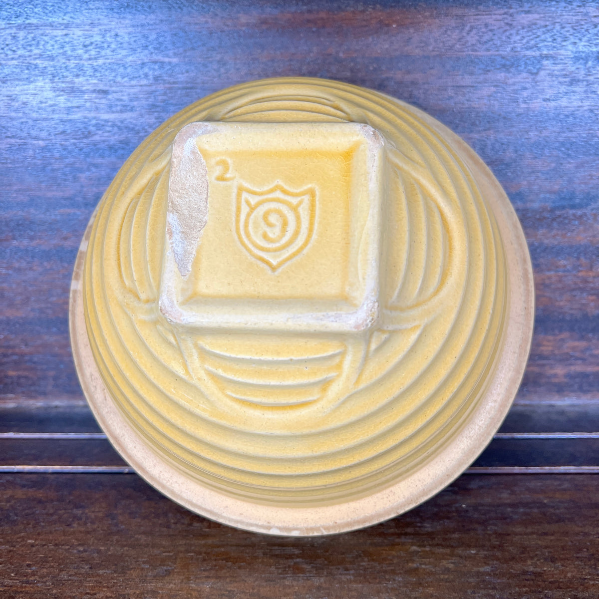 A top-down view of a round, light yellow pottery or small bowl with concentric circular ridges and a square pedestal base featuring an embossed shield-like emblem with the number 9. The bowl is placed on a dark wooden surface.