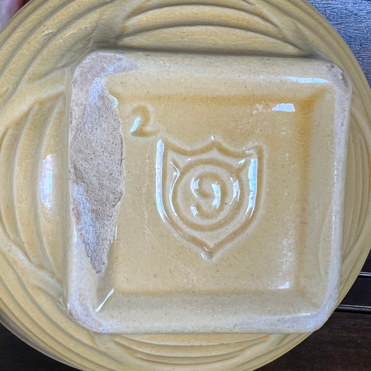 Close-up of a square ceramic dish or plate with a raised shield-like emblem in the center, shown on a matching round plate. The glaze is a warm pale yellow-beige with a glossy finish and a visible chip on the left side.