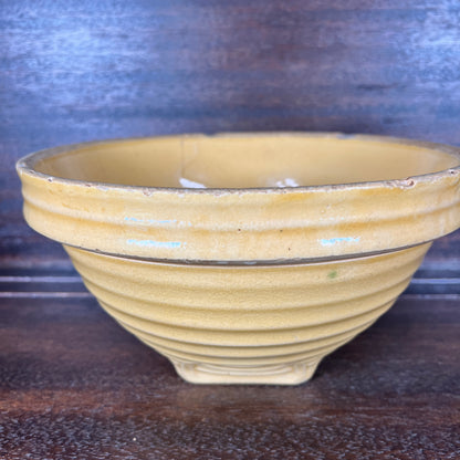 A vintage yellow mixing bowl with a ribbed exterior and a small footed base, shown on a dark wooden surface against a dark blue textured background.