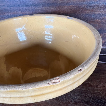 A close-up of a round ceramic bowl with a glossy mustard-yellow glazed interior and a rustic, unglazed brown rim, shown on a dark wooden surface.