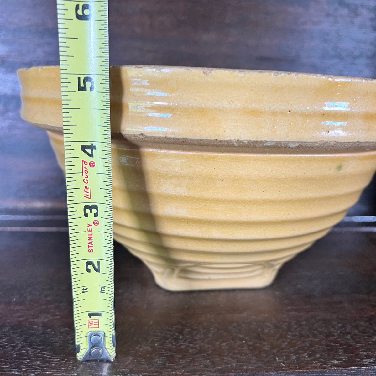 A yellow bowl with a ribbed exterior and a brown rim, shown next to a Stanley tape measure on a dark wooden surface.