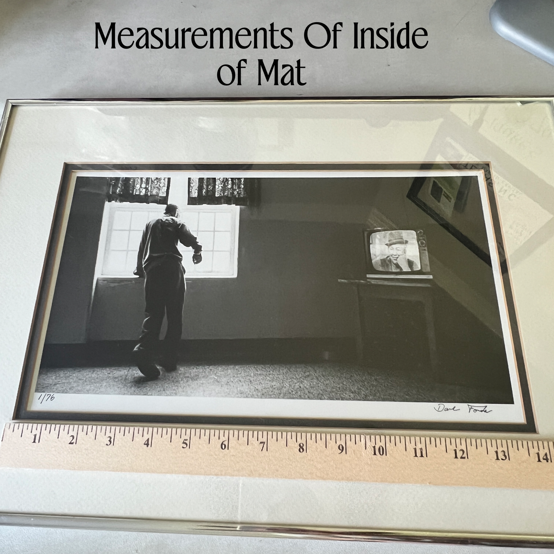 A framed black-and-white art print titled 'Measurements of Inside of Mat' shown with a measuring tape along the bottom to indicate scale. The print depicts a person standing in a dimly lit room near a window, with a small television on a table in the background.