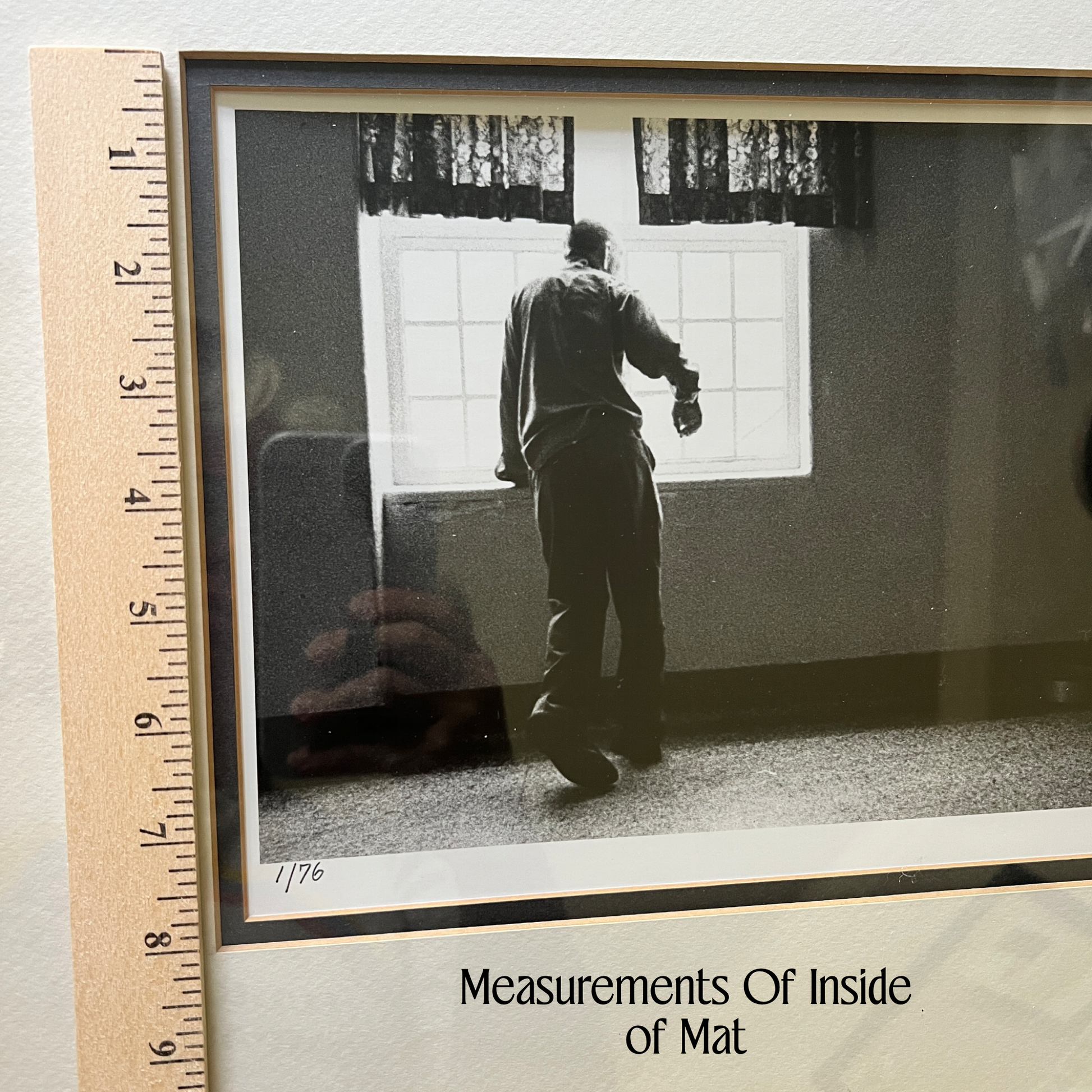 A framed black-and-white photograph showing a person standing in a room near a window, placed on a light surface with a wooden ruler along the left edge for scale. Text at the bottom reads 'Measurements Of Inside of Mat.