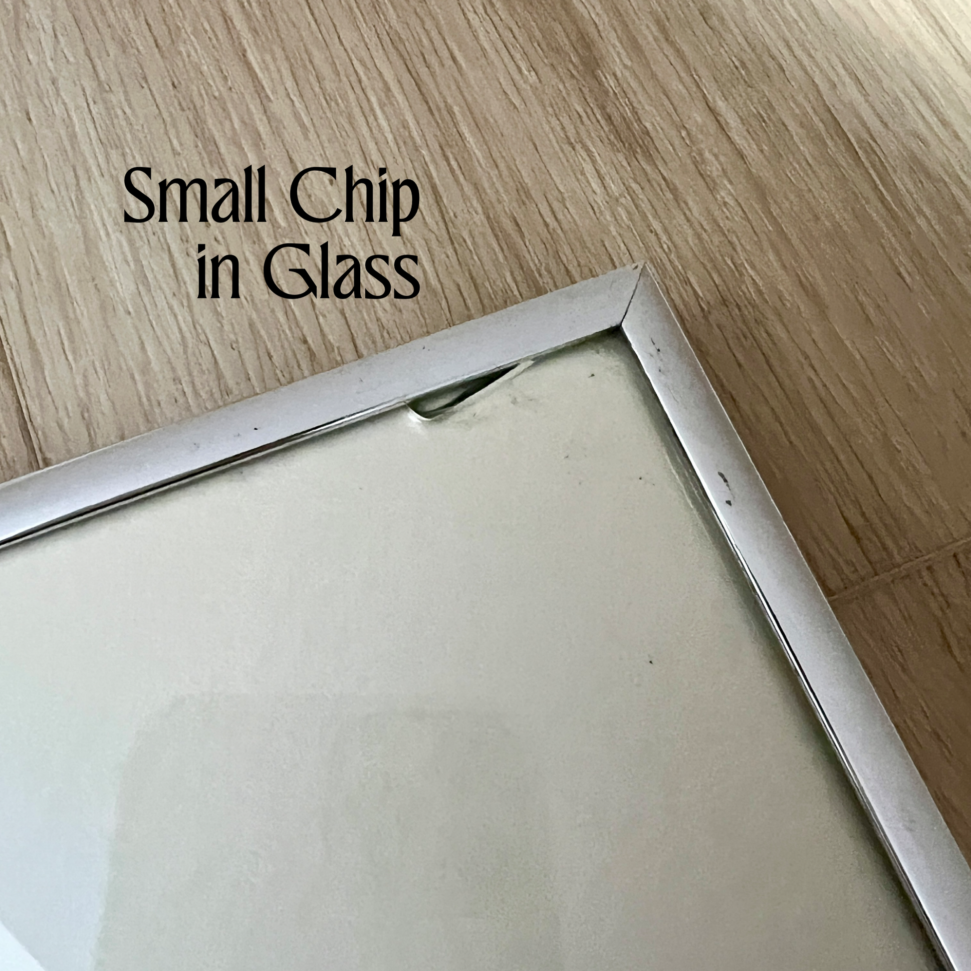 Close-up of the corner of a picture frame with a silver metal border and clear glass, placed on a light wood floor. Black text in the upper left reads 'Small Chip in Glass'.