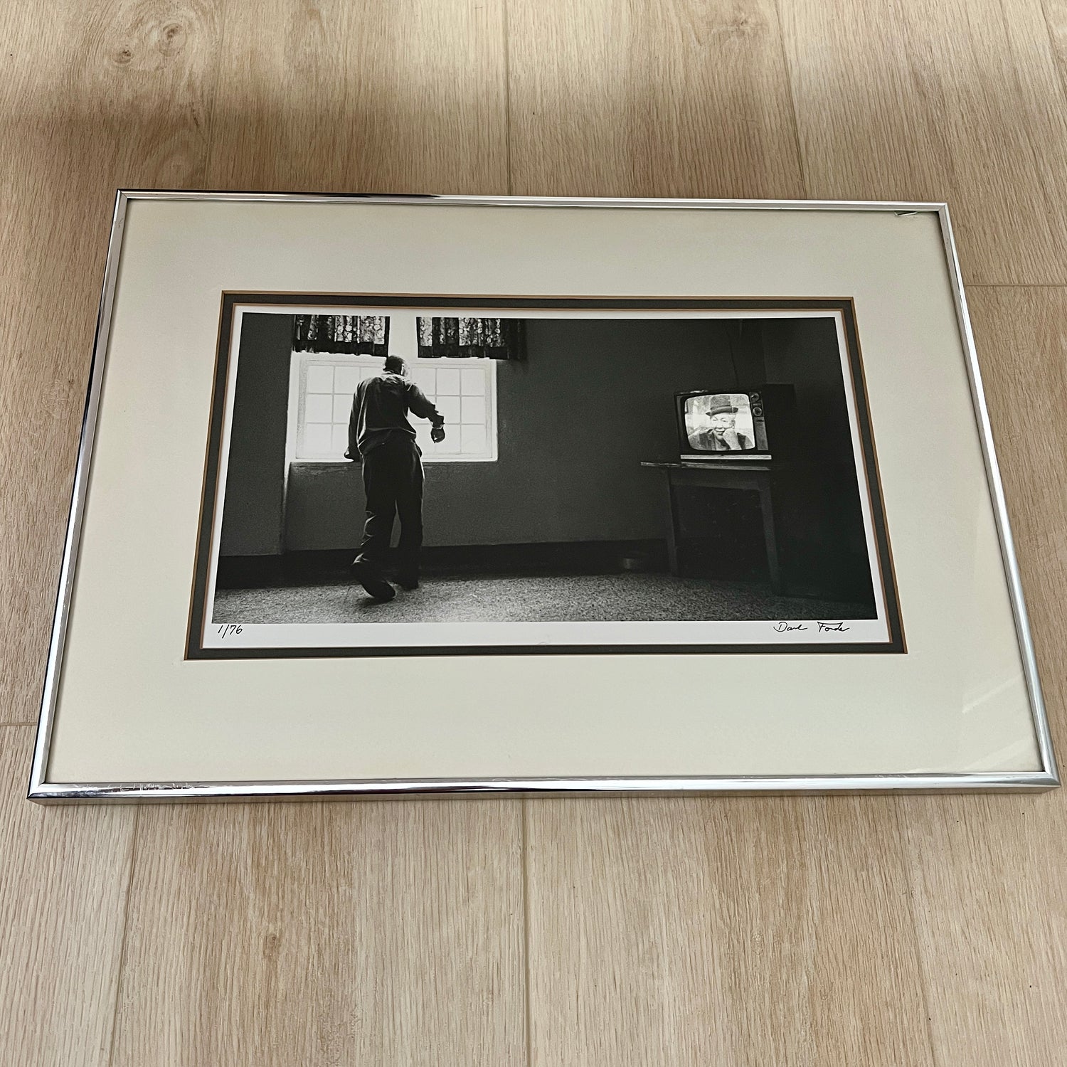 A framed black and white art print featuring a vintage photograph of a man standing in a room next to a television, displayed on a light wood floor.