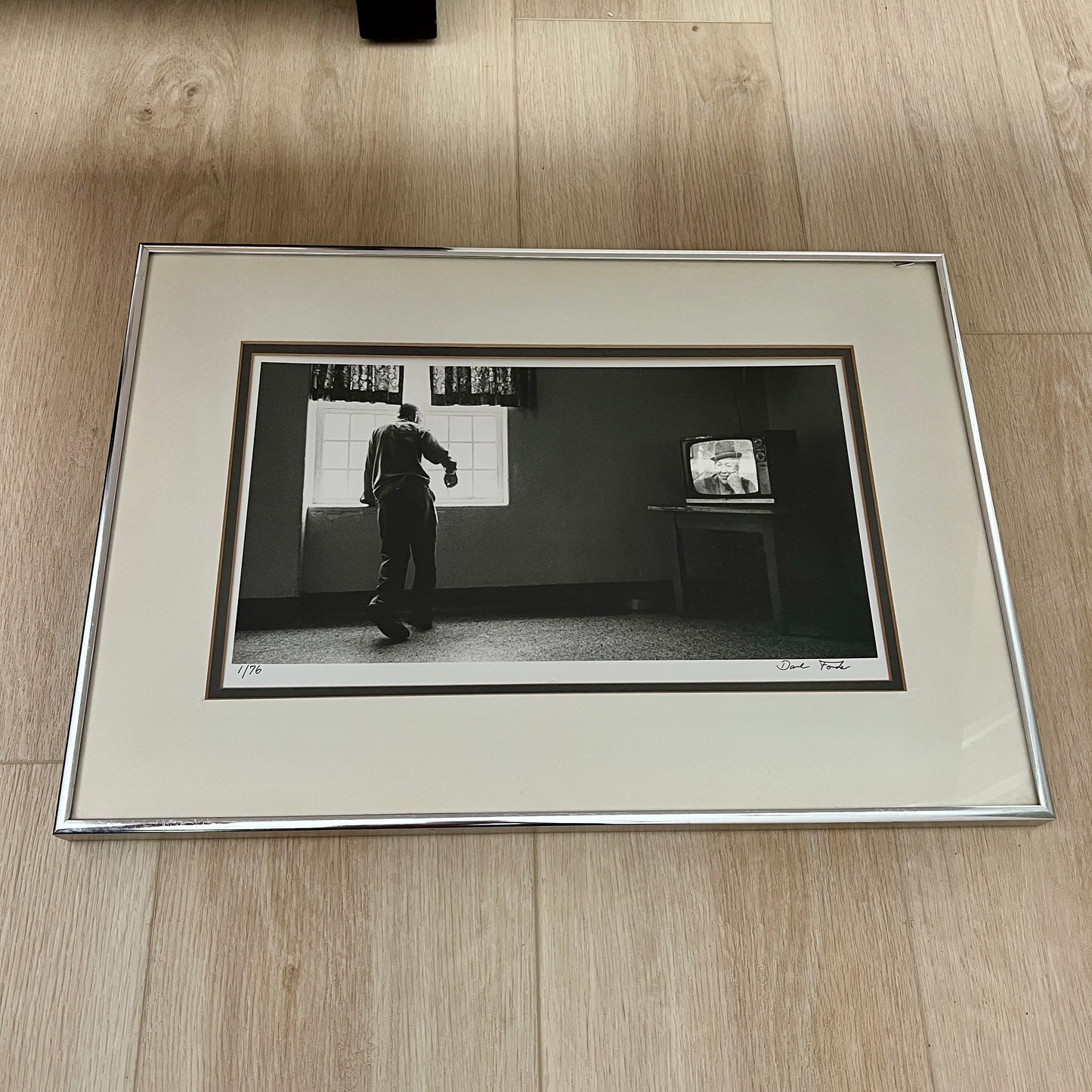 A framed black and white art print featuring a photographer's scene of a man walking past a vintage television set in a dark room, displayed on a light wood floor.