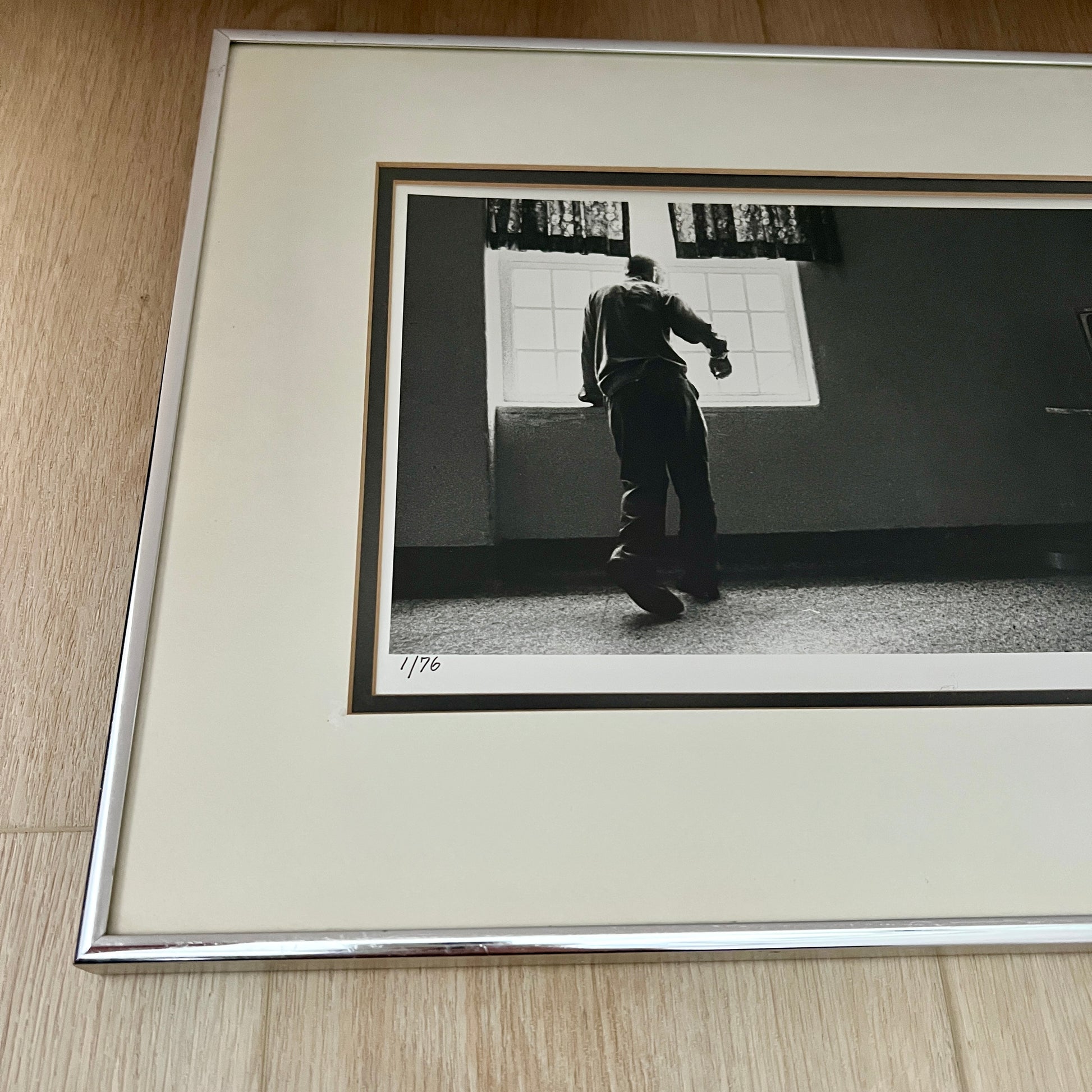 A framed black and white art print featuring a photograph of a man standing in a room, mounted with a wide cream mat and a polished silver metal frame, resting on a light wood floor.