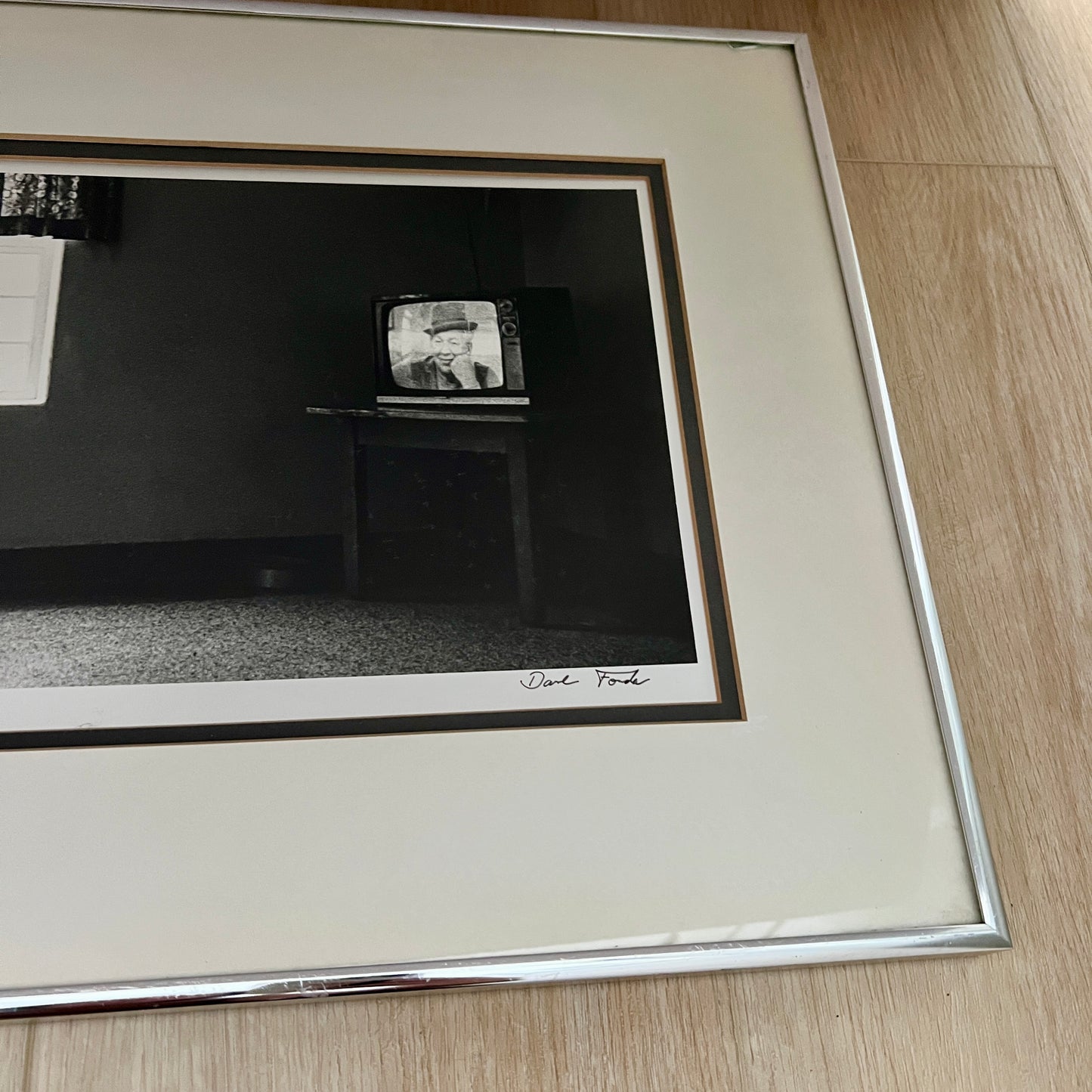A framed black and white art print featuring a photograph of a vintage television set on a table, with a wide cream mat and a silver metallic frame, placed on a light wood floor.