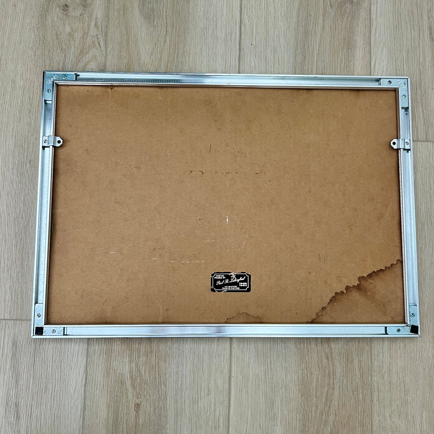 The back of a picture frame with a silver metal border and a brown cardboard backing, placed on a light wood floor. With water damage on cardboard