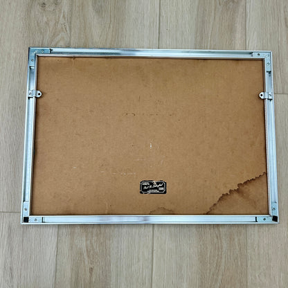 The back of a picture frame with a silver metal border and a brown cardboard backing, placed on a light wood floor. With water damage on cardboard