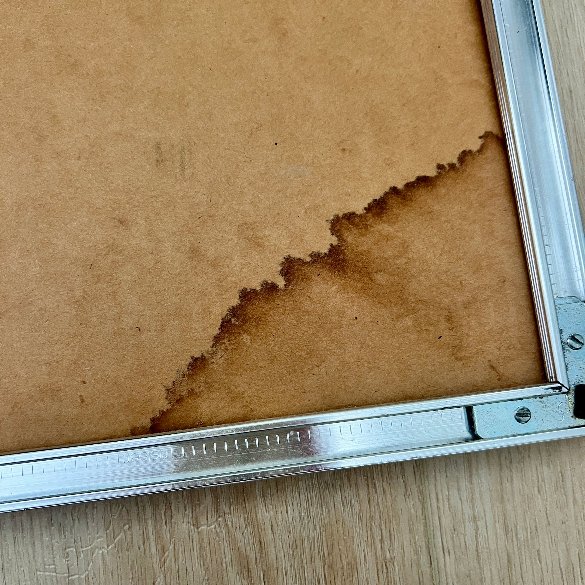 Close-up of the back side of a square picture frame with a brown mounting board and a silver metal frame corner on a light wood surface. Shows water staining in corner