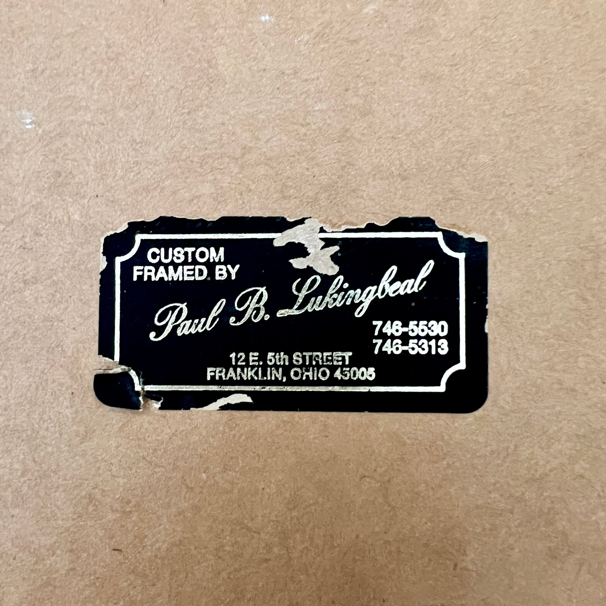 A black custom framed art label with white text reading 'Custom Framed by Paul R. Luingbeal' on a brown kraft paper background.
