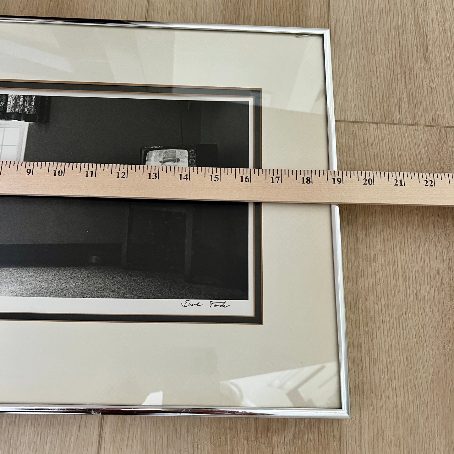 A framed black and white art print with a cream mat and silver frame, shown on a light wood floor with a wooden ruler laid across the mat to indicate size.