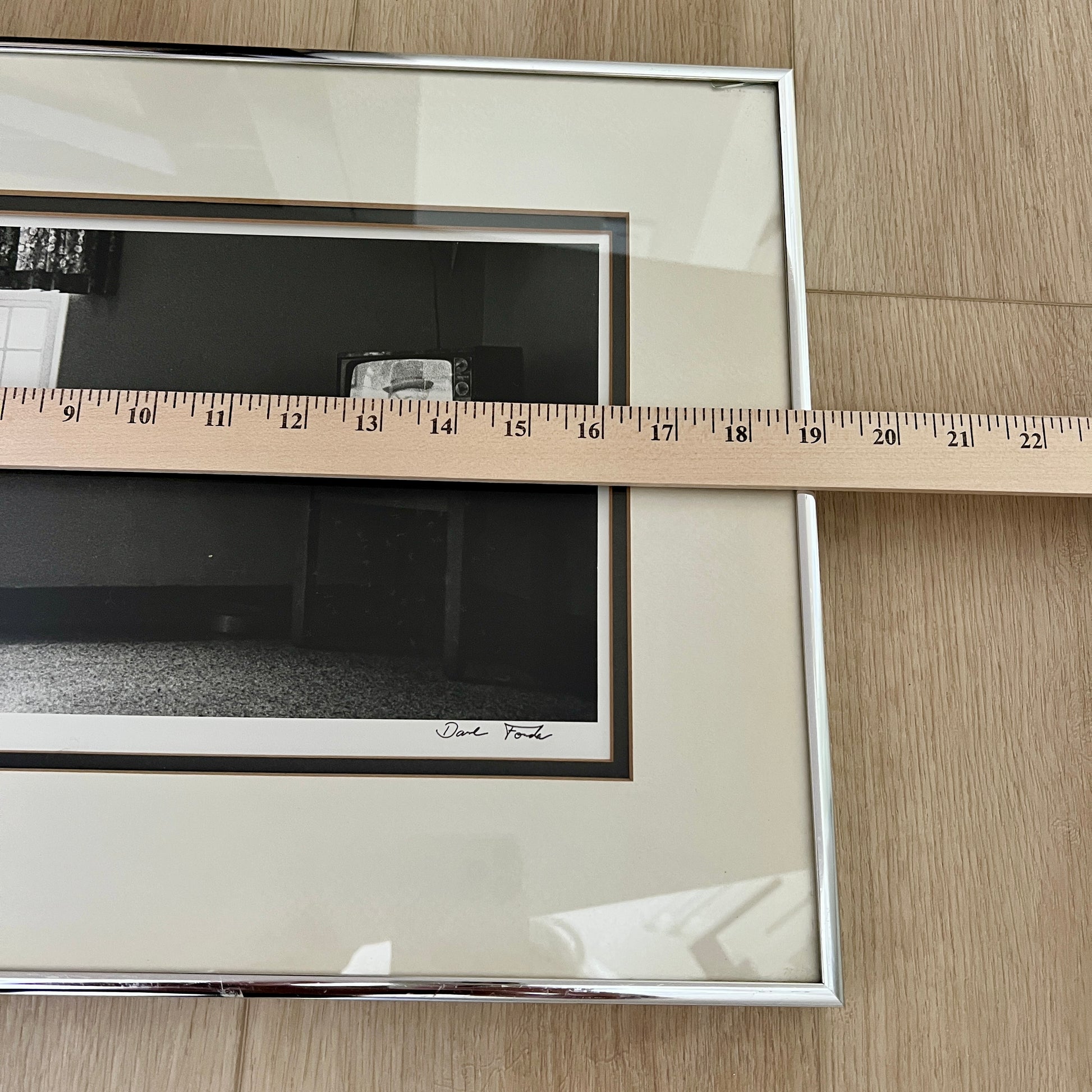 A framed black and white art print with a cream mat and silver frame, shown on a light wood floor with a wooden ruler laid across the mat to indicate size.