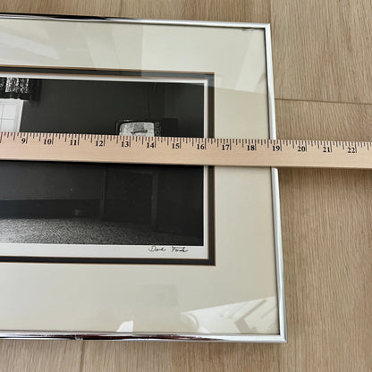 A framed black and white art print with a cream mat and silver frame, shown on a light wood floor with a wooden ruler laid across the mat to indicate size.
