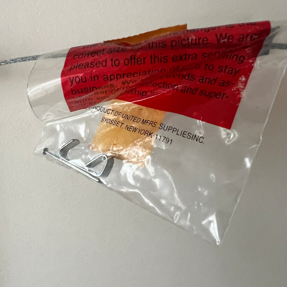 A clear plastic bag containing a small metal hook or clip, with a red printed label and partial text visible on the bag, photographed against a plain light wall.