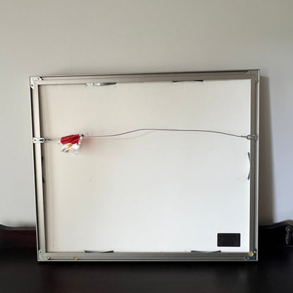 The back side of a square framed wall art piece or mirror, showing a silver metal frame, mounting hardware, a wire across the center, and a small black label in the bottom right corner, photographed against a plain indoor wall.