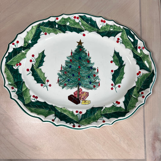 An oval white ceramic Christmas serving platter with a scalloped edge, decorated with a green holly leaf border and red berries, featuring a central illustration of a decorated Christmas tree with presents.