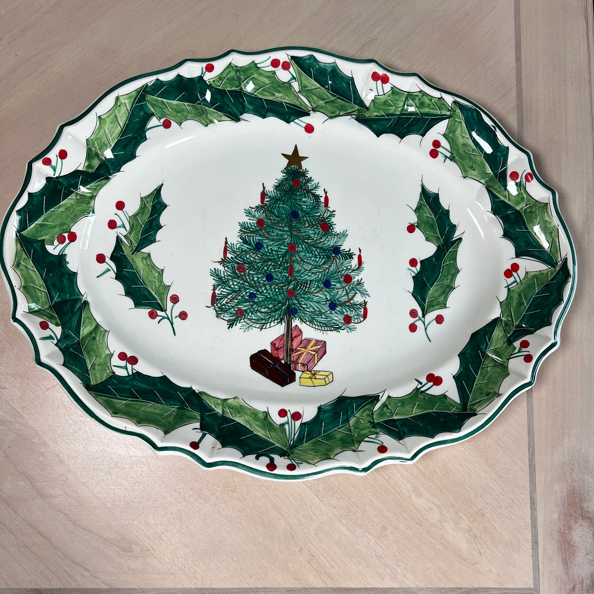 An oval white ceramic Christmas serving platter with a green holly and red berry border, featuring a decorated Christmas tree and wrapped gifts in the center, shown on a light wood surface.